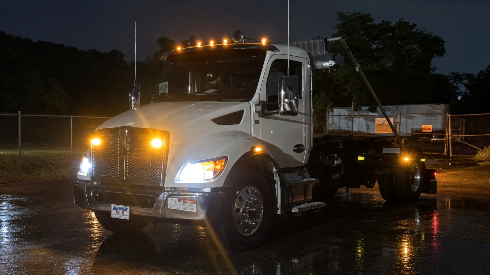 Kenworth hooklift truck upfitted by Auto Action Technologies with amber roof-mounted warning lights and grille strobes lit up at night on a wet lot